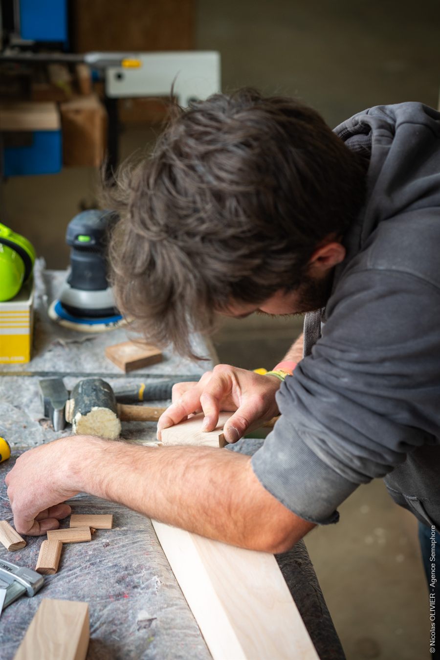 Une personne ajuste une pièce de bois sur un établi, entourée d'outils à main.
